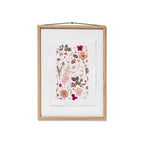 A framed botanical art print featuring pressed flowers and leaves in soft pink, red, beige, and green tones, set in an oak frame with a white base and hanging cord against a white background.
