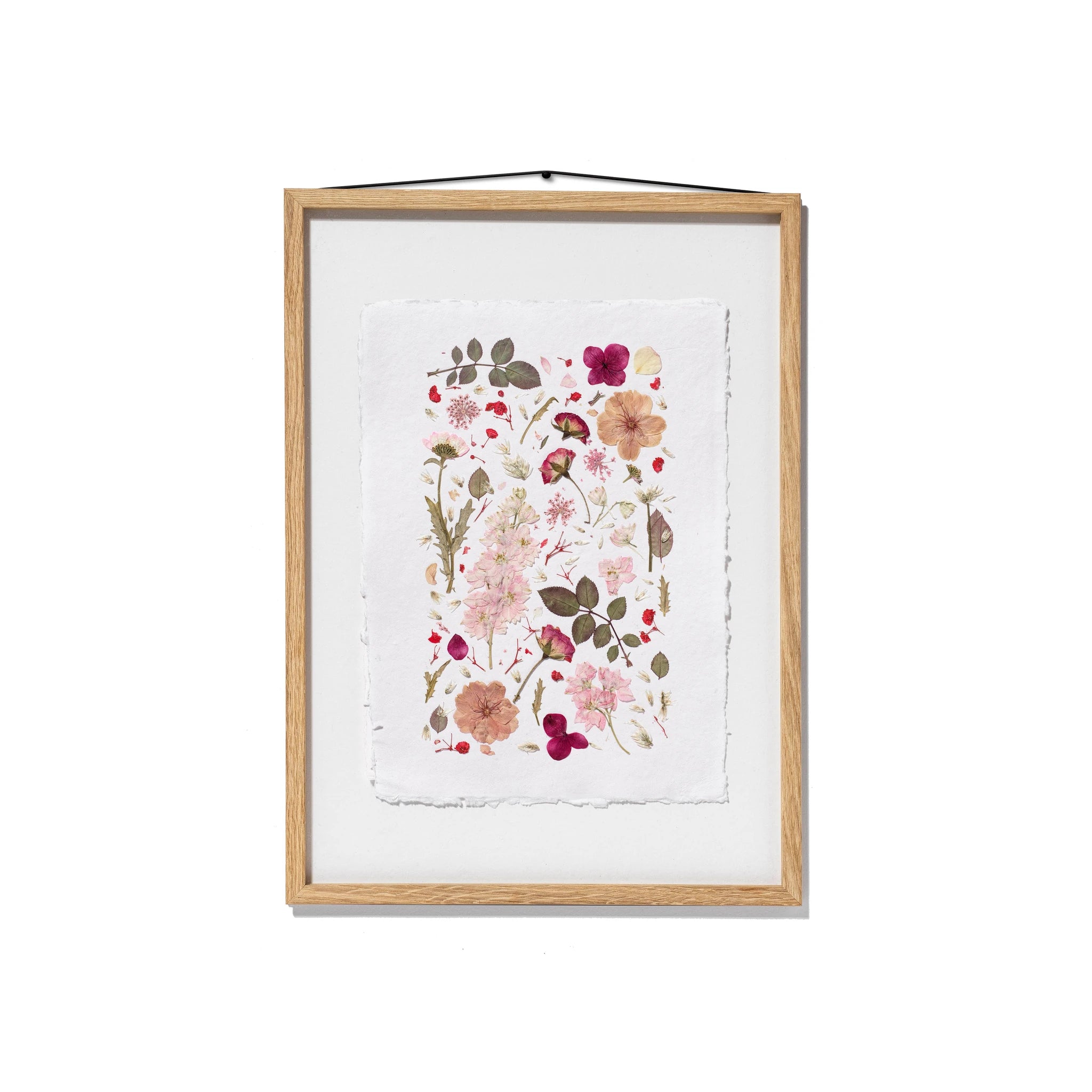 A framed botanical art print featuring pressed flowers and leaves in soft pink, red, beige, and green tones, set in an oak frame with a white base and hanging cord against a white background.