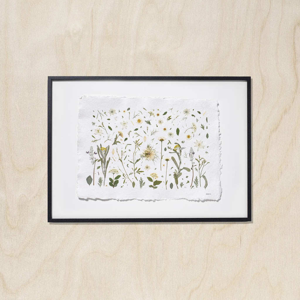 A framed botanical art featuring delicate flowers on a textured white sheet of paper, mounted with a white background and a black frame against a light wood background.