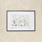 A framed botanical art featuring delicate flowers on a textured white sheet of paper, mounted with a white background and a black frame against a light wood background.