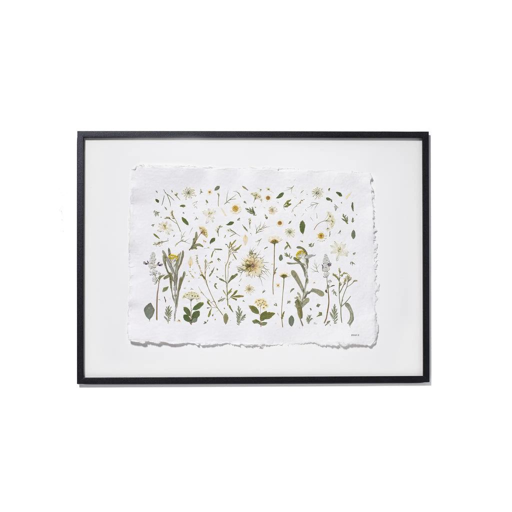 A framed botanical art featuring delicate pressed flowers and green leaves on a textured white paper background, enclosed in a black frame with a white backdrop.